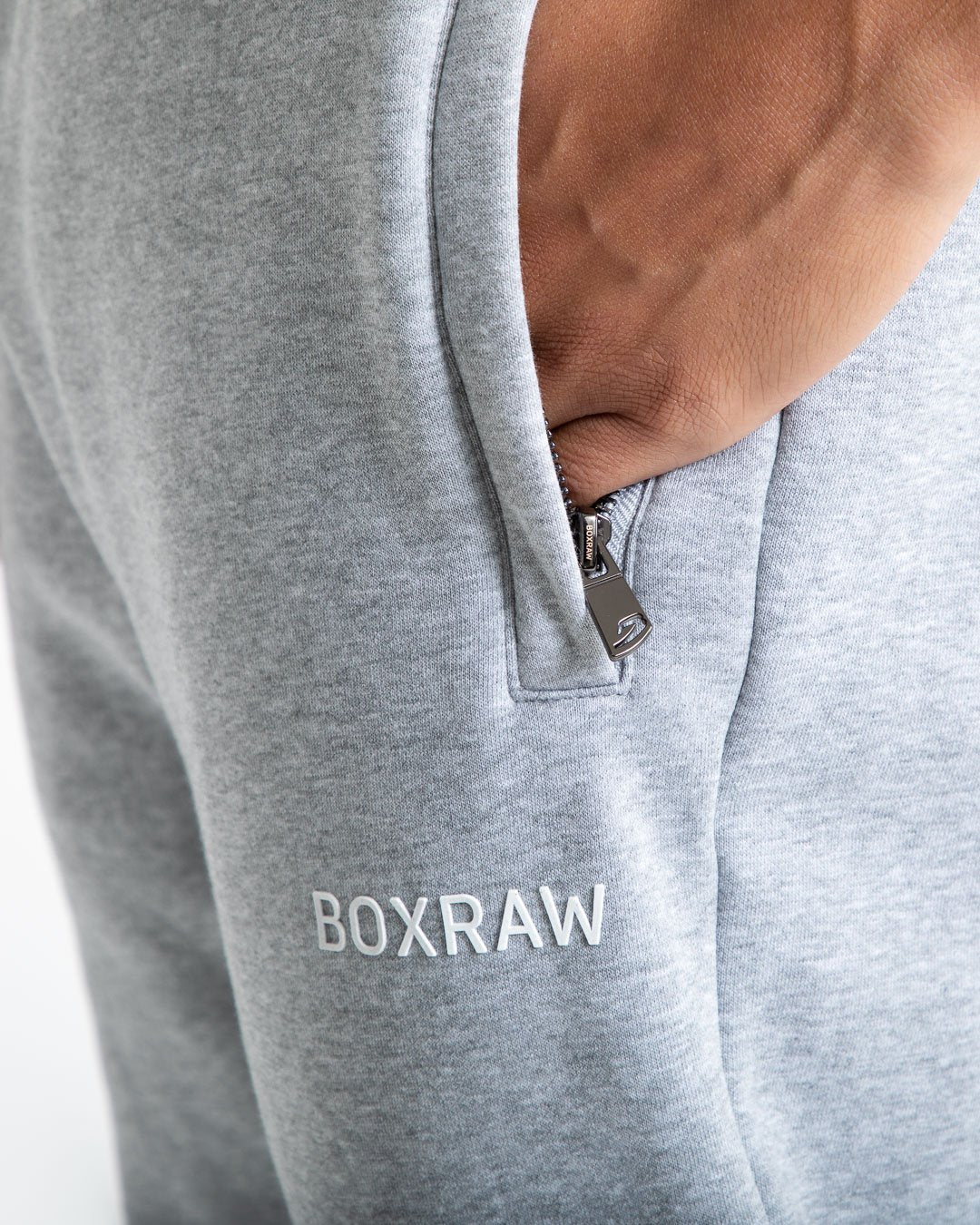 Man in relaxed fit grey tracksuit bottoms with a white 3D boxraw logo as well as side zipped pockets made from a cotton/poly blend.