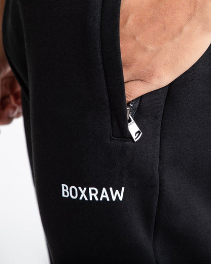 Man in relaxed fit black tracksuit bottoms with a white 3D boxraw logo as well as side zipped pockets made from a cotton/poly blend.