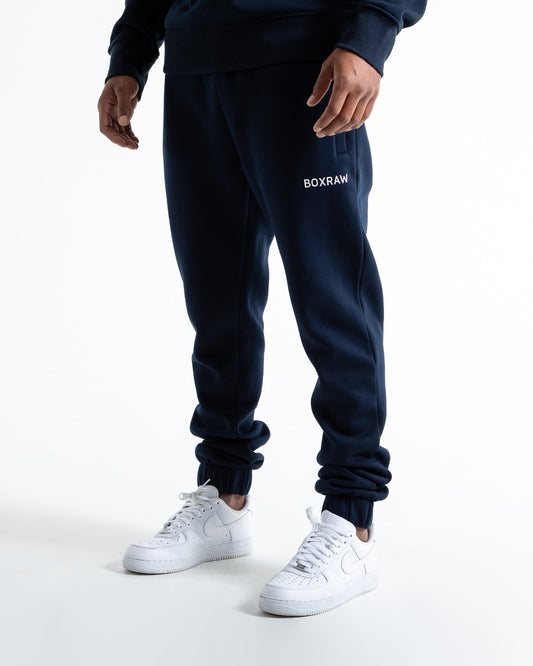 Man in relaxed fit navy tracksuit bottoms with a white 3D boxraw logo as well as side zipped pockets made from a cotton/poly blend.