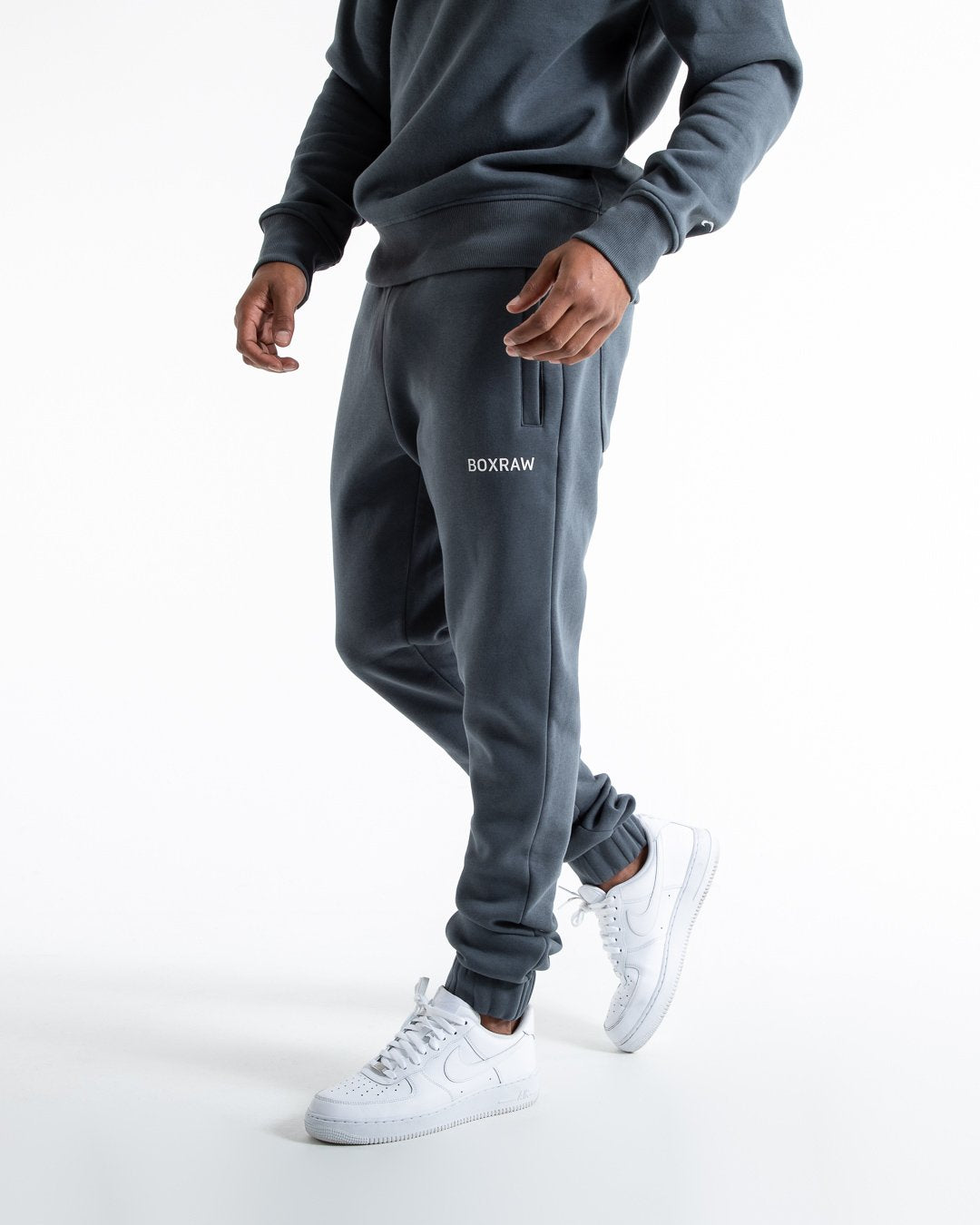 Man in relaxed fit charcoal tracksuit bottoms with a white 3D boxraw logo as well as side zipped pockets made from a cotton/poly blend.