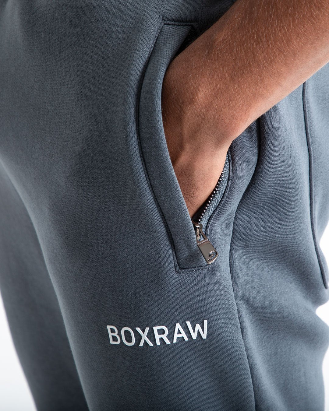 Man in relaxed fit charcoal tracksuit bottoms with a white 3D boxraw logo as well as side zipped pockets made from a cotton/poly blend.