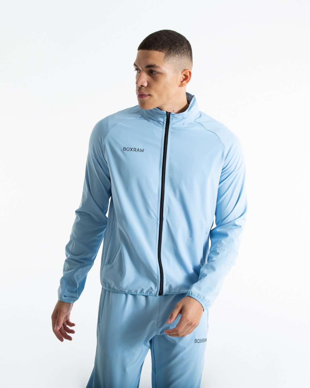 Man in baby blue zipped lightweight tracksuit jacket with a hidden zipped hoodie with black boxraw logo made from polyester.