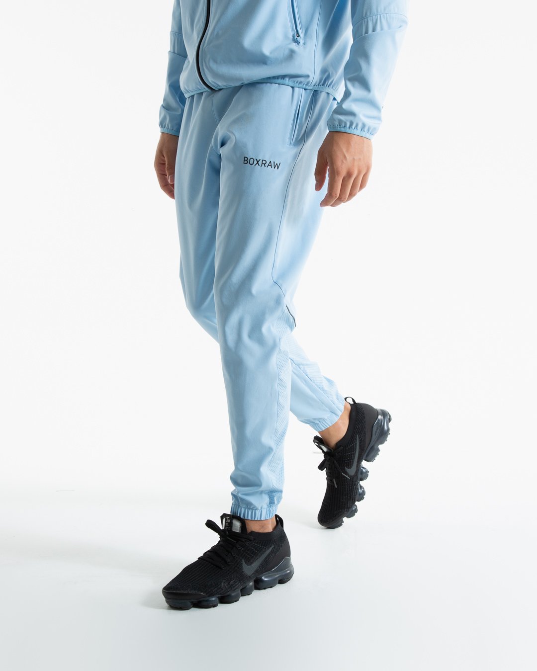 Man in baby blue lightweight tracksuit bottoms with mesh/perforated ventilation panels on the legs with black boxraw logo