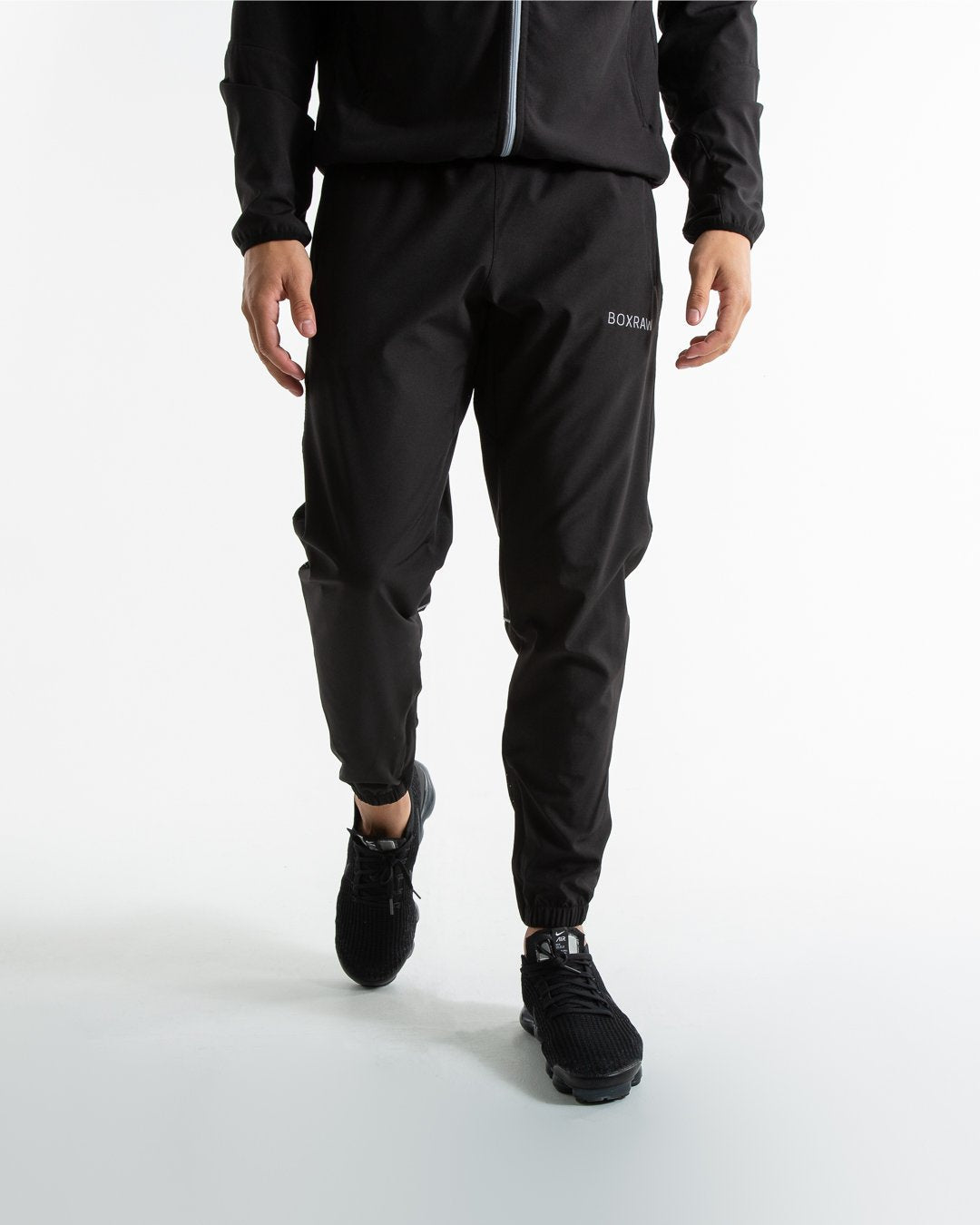 Man in black lightweight tracksuit bottoms with mesh/perforated ventilation panels on the legs with white boxraw logo.