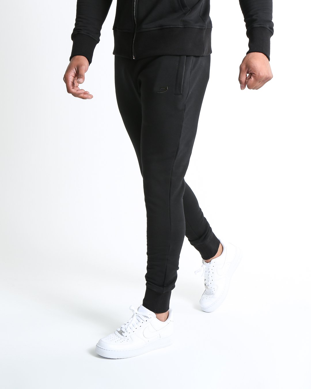 Man in black slim fit tracksuit bottoms with metal strike logo and zipped side pockets